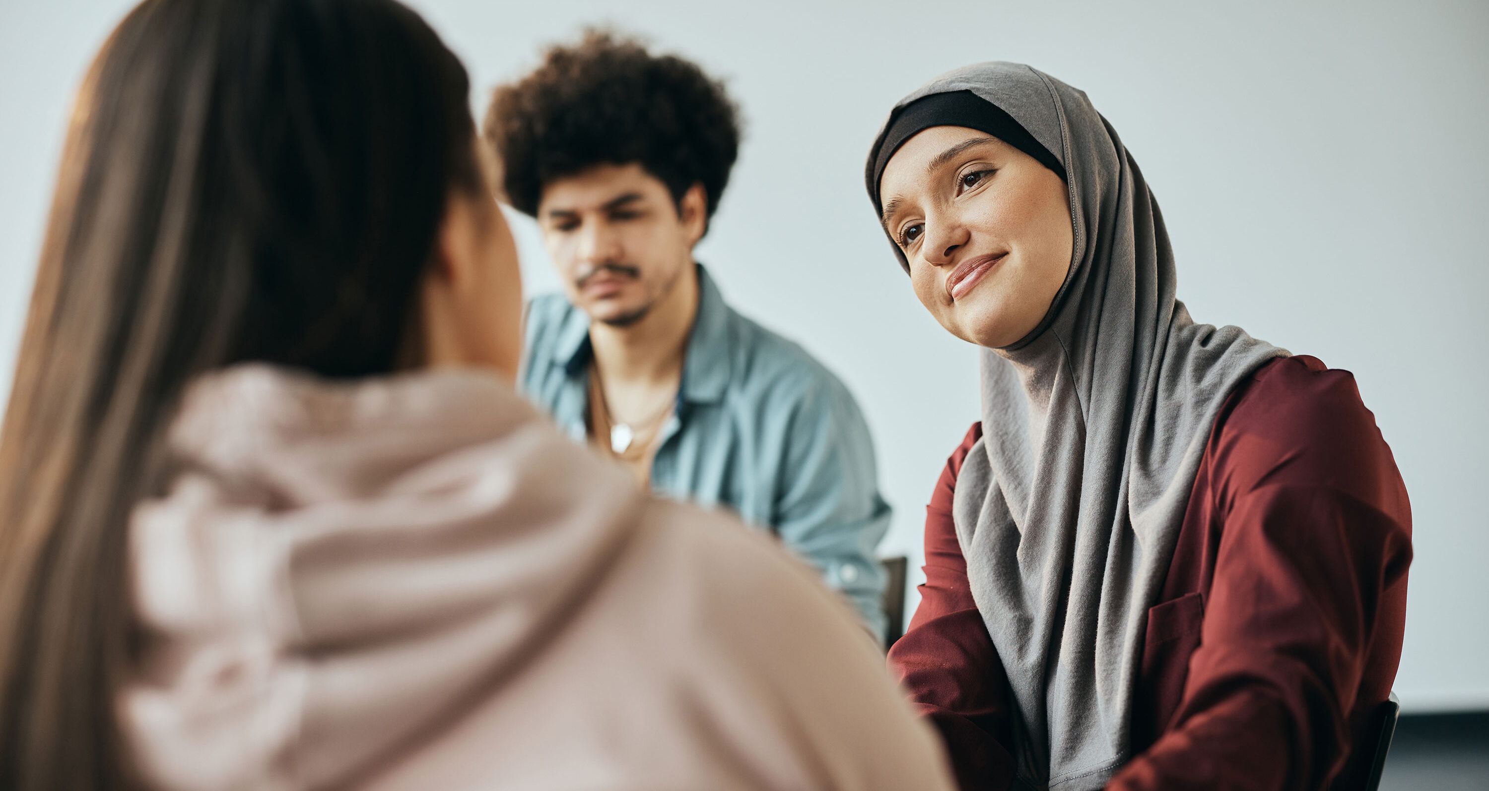 Im Fokus steht eine Frau mit Kopftuch, die sich der Person neben ihr freundlich zuwendet. Im Hintergrund ist eine weitere Person zu sehen. Sie sitzen im Halbkreis und könnten an einer Fortbildung teilnehmen.